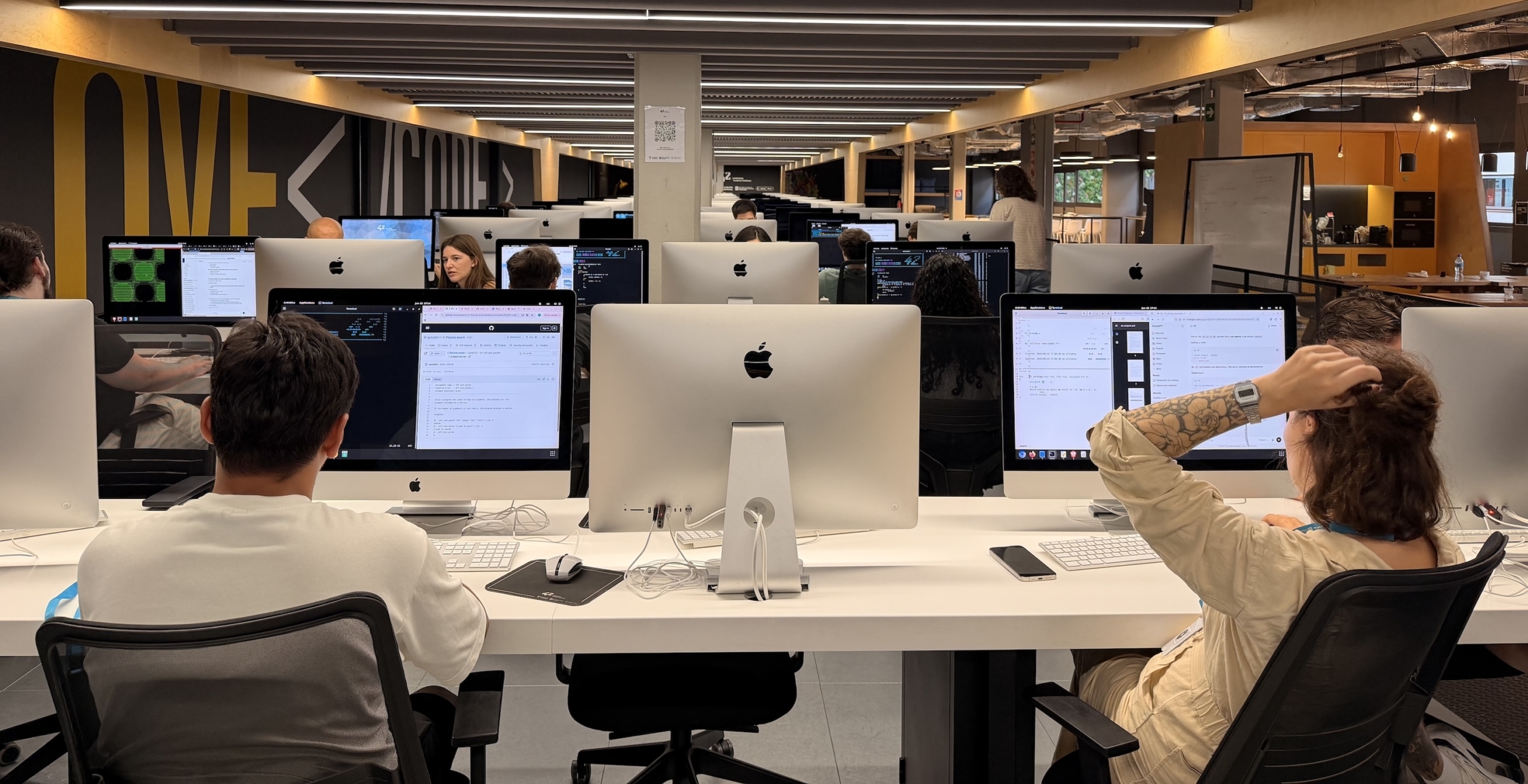 Interior del campus de programación 42 Barcelona con filas de mesas y ordenadores preparados para programar; en la pared se lee “We love code”.