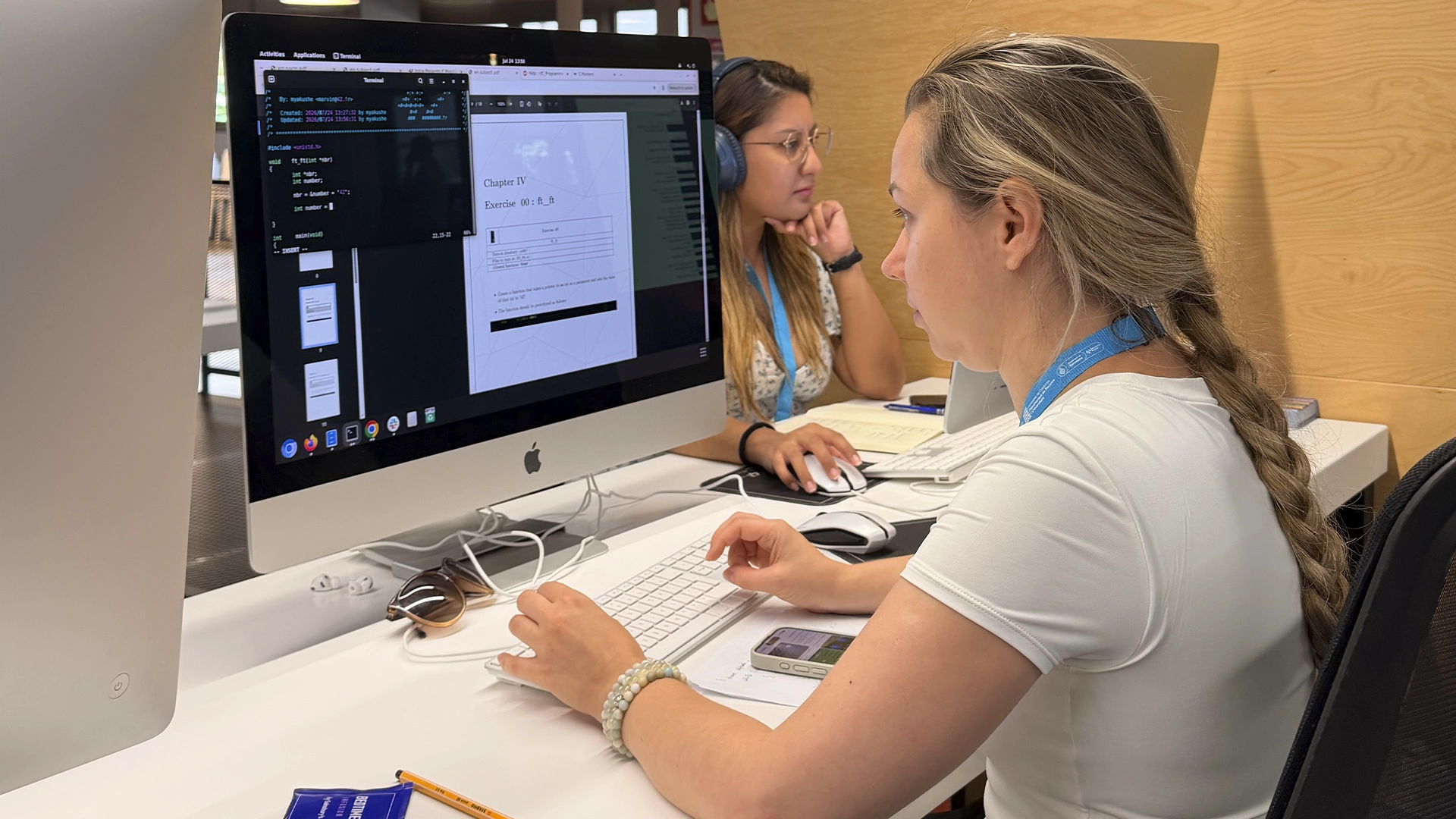 Two women collaborate at a shared desk with large desktop computers, one typing while the other talks, in a bright office setting.