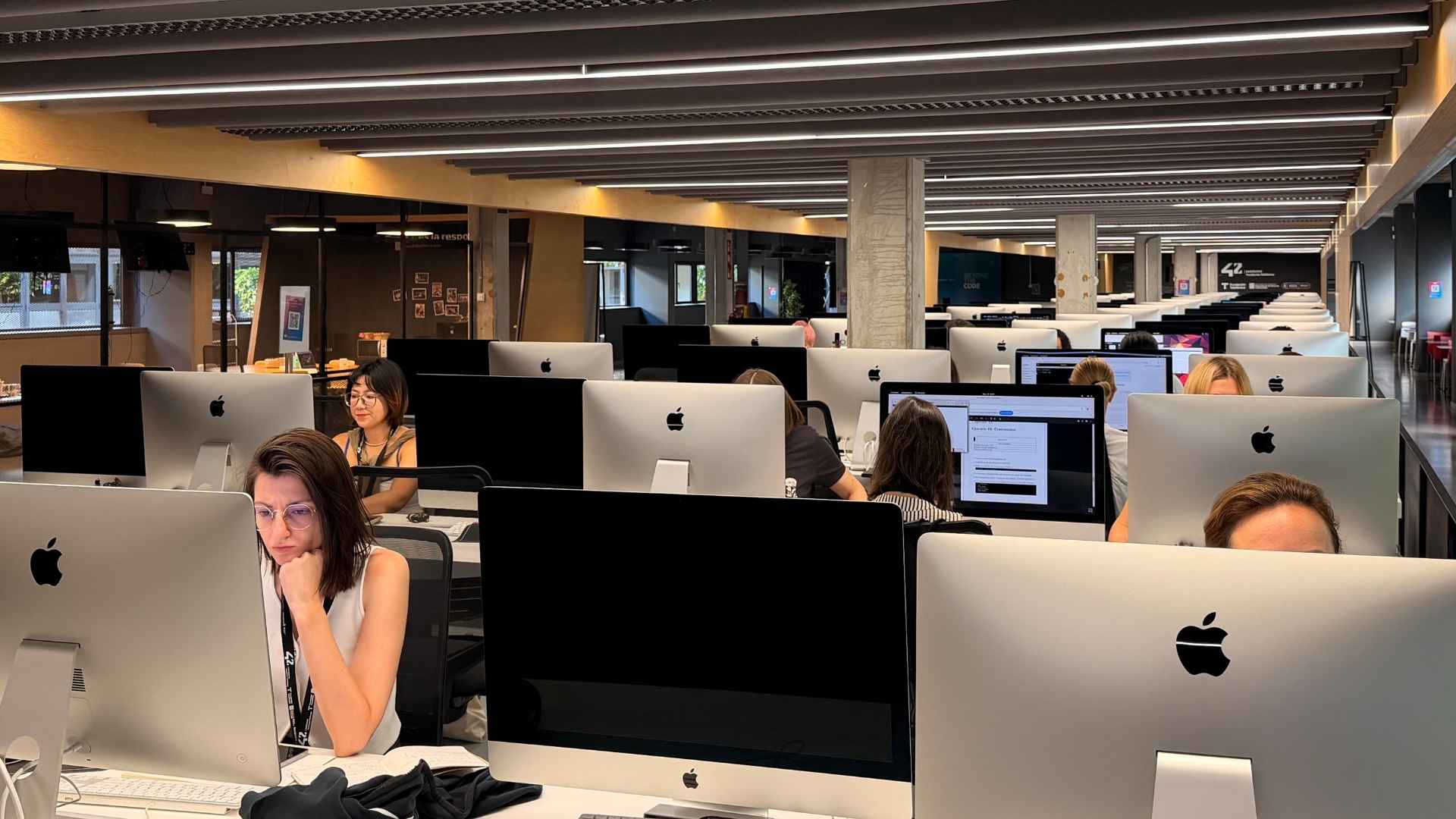 Open-plan office with rows of Apple iMacs and workers at desks under modern ceiling lights.